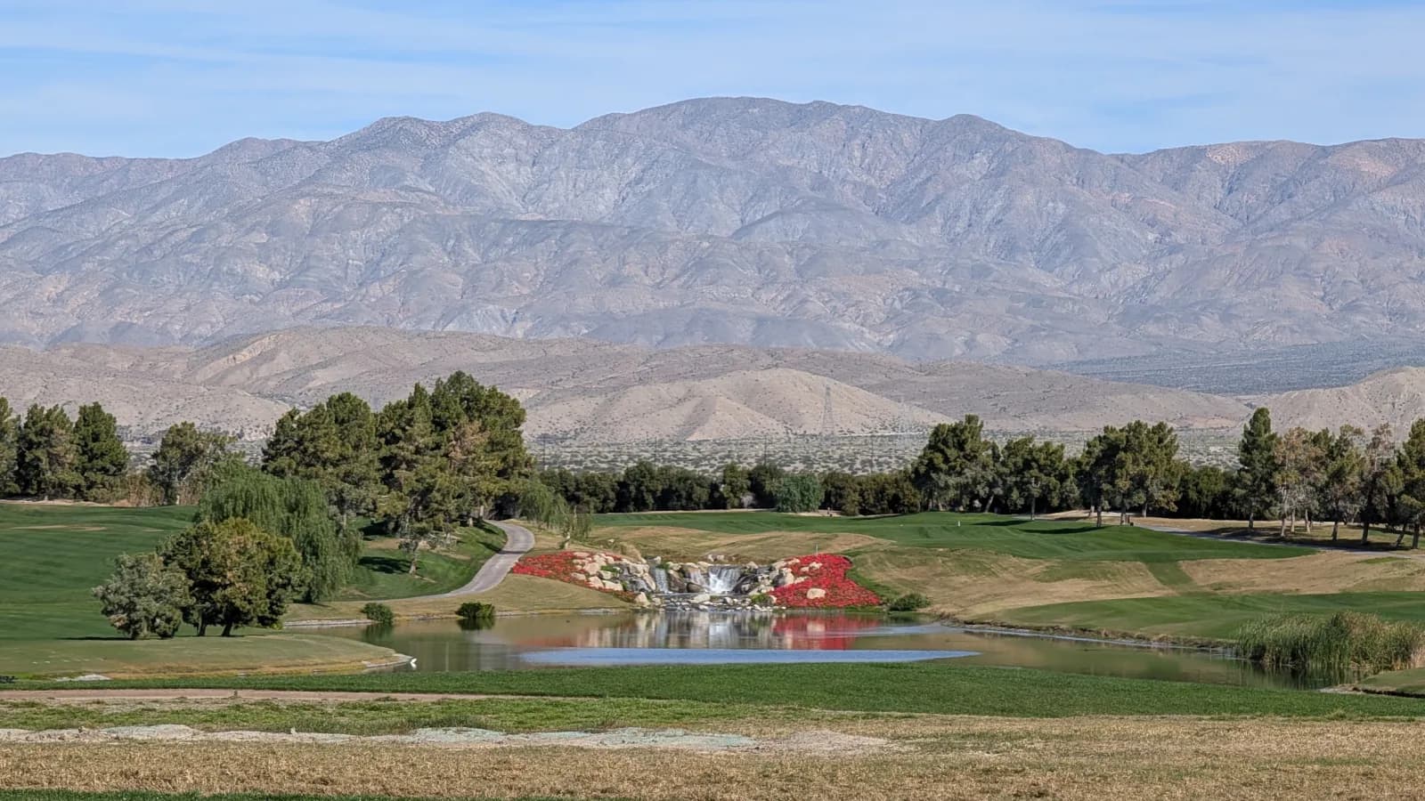 A pond and rock waterfall banked with red flowers at Classic Club, a cart path curving past behind it and a long mountain ridge filling the horizon.