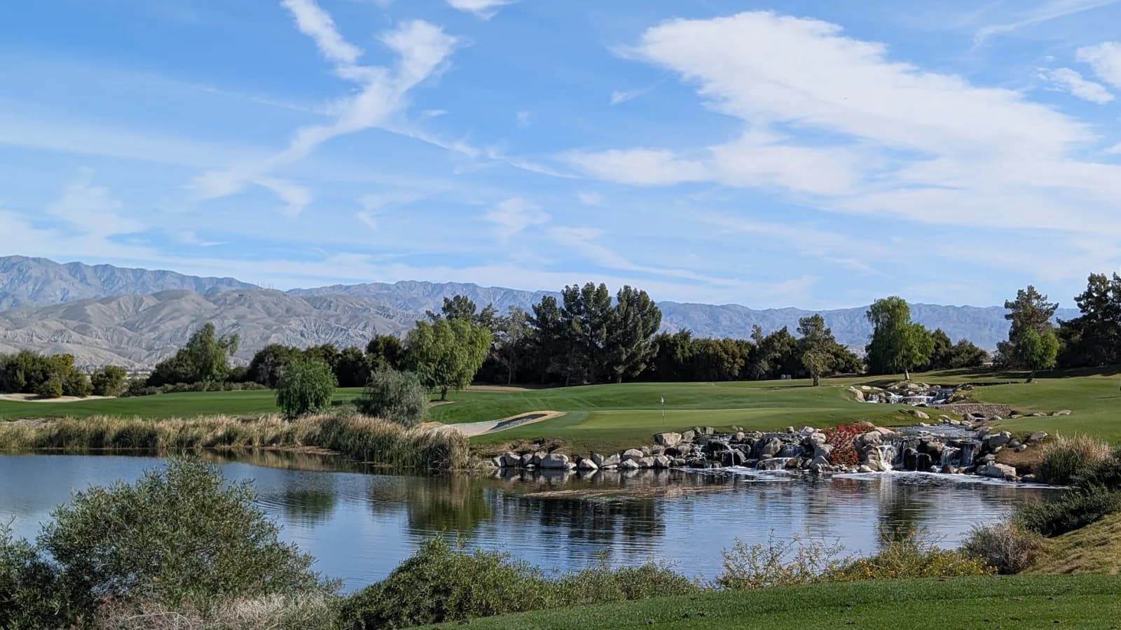 The par-3 12th at Classic Club seen from across the water, the green and flag on the far bank with a rock waterfall and red flowers to the right.