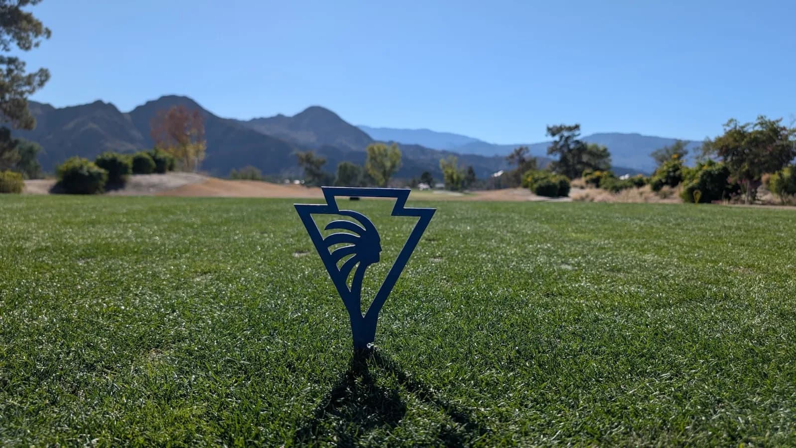 A blue metal Indian Wells tee marker planted in the turf in close focus, with the fairway and mountains soft behind it.