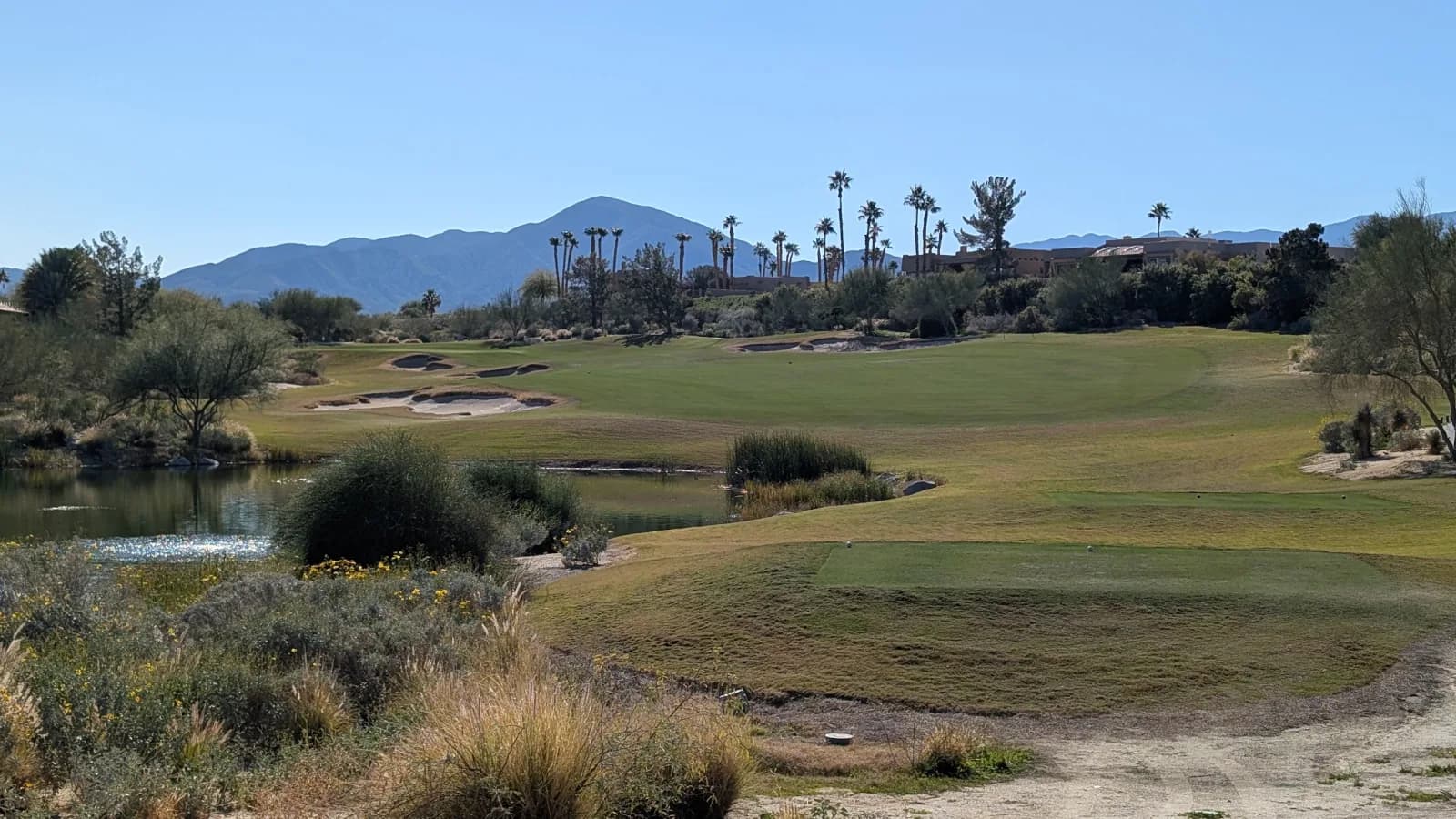 A green at Rams Hill ringed by bunkers, seen across a reed-lined pond, with palms, adobe-style buildings, and a mountain ridge behind.