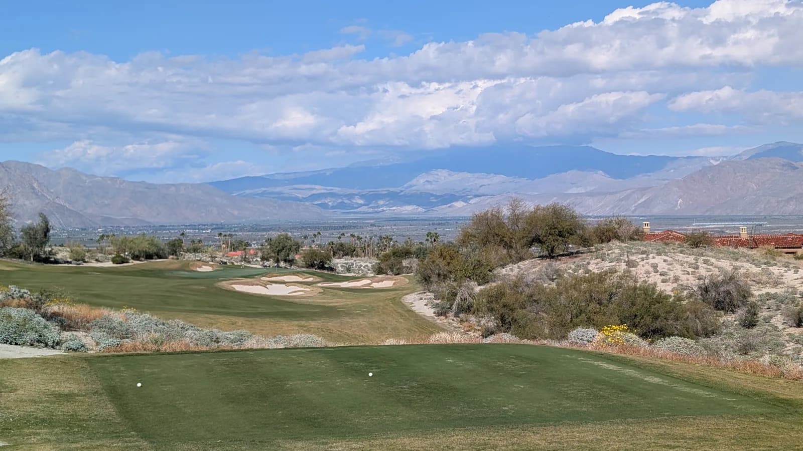 The view from an elevated Rams Hill tee box, two balls teed up in the foreground, a cluster of bunkers guarding the fairway below, and a wide desert valley running out to distant mountains.