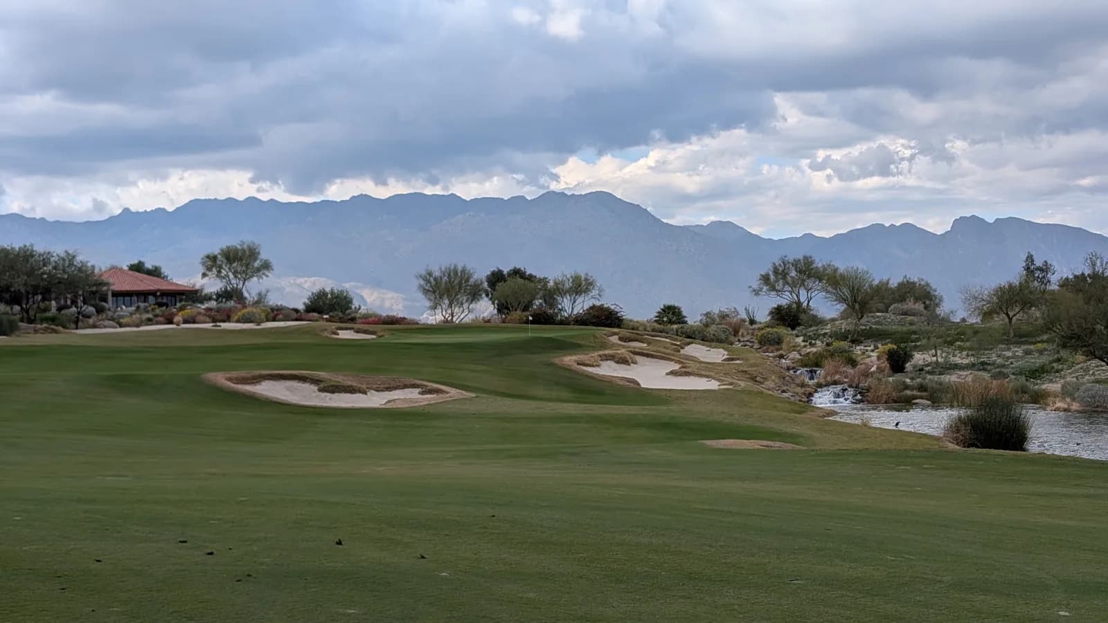 A Rams Hill fairway running up to a flagged green flanked by large bunkers, with a rocky stream spilling into a pond on the right and a tile-roofed clubhouse under heavy cloud on the left.