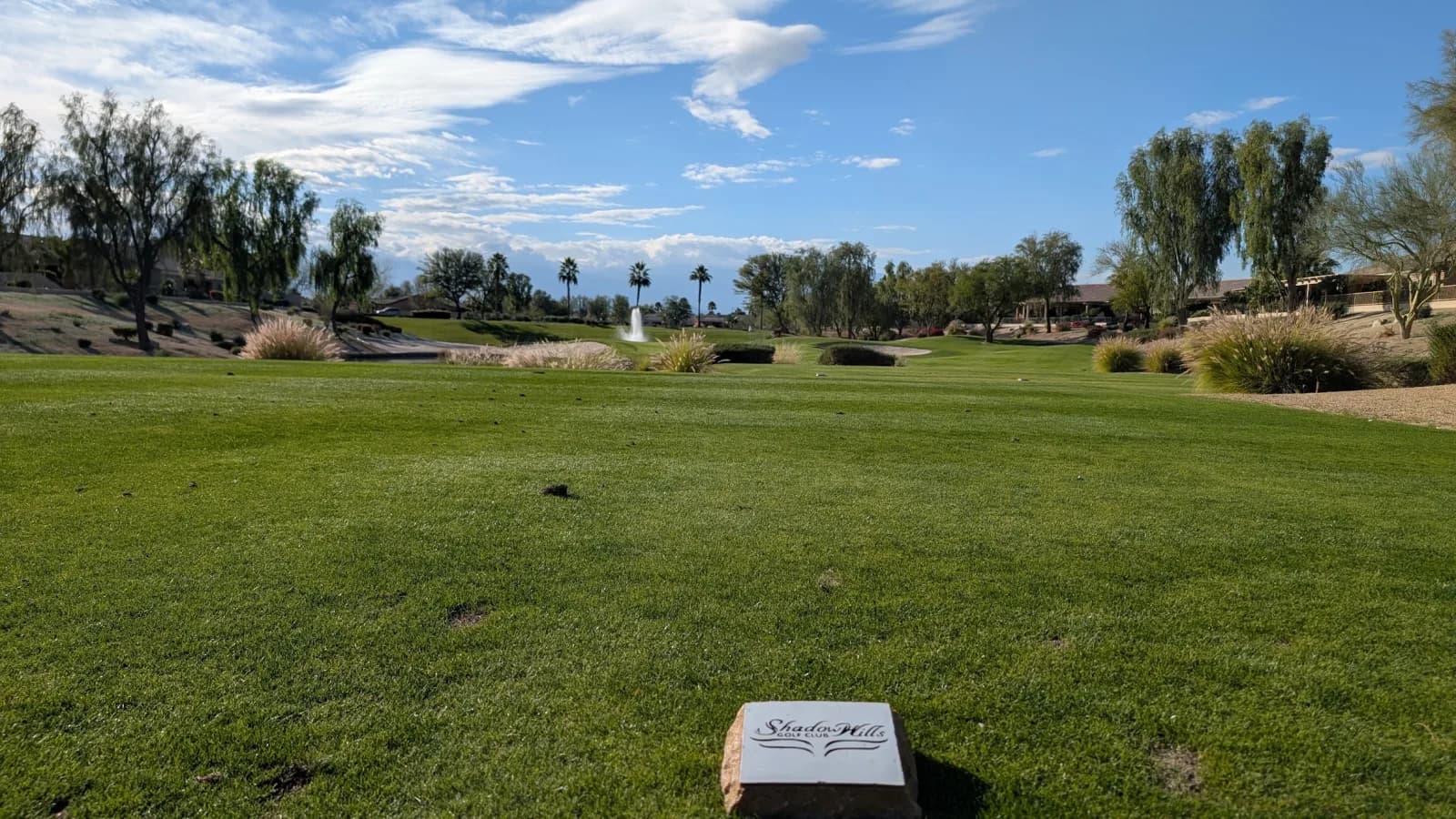 A Shadow Hills Golf Club tee marker set in the turf at the front of a tee box, looking down a wide green fairway toward a fountain in a pond, with willows, palms, and desert landscaping beyond.
