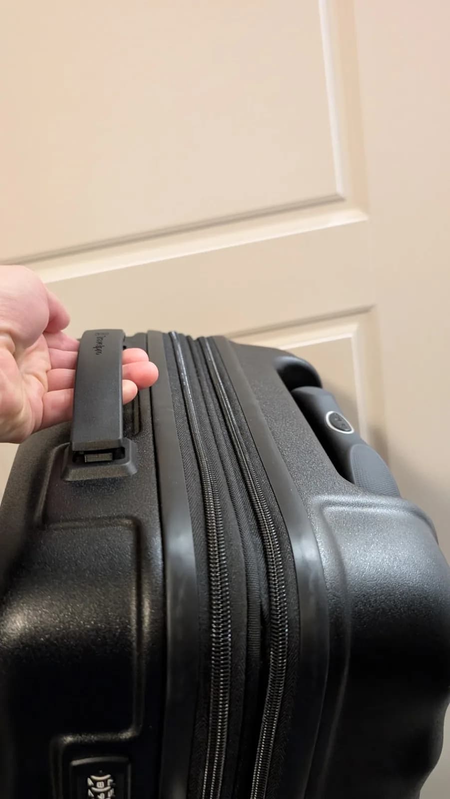 A hand lifting the bag by the top carry handle alone, the case hanging below it with the telescoping handle visible along the far edge.